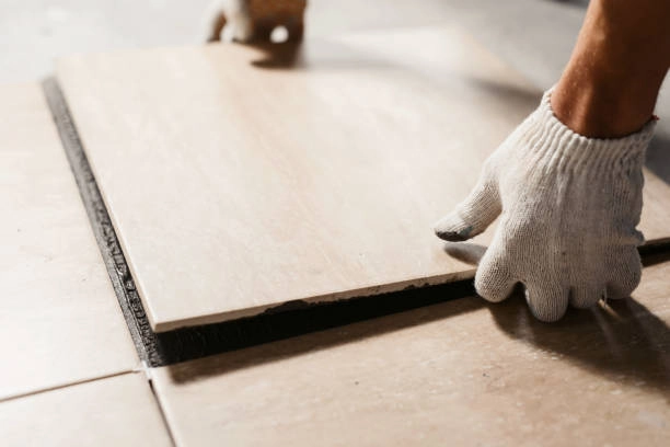 Gloved worker placing large ceramic tile onto adhesive flooring base. -flooring installation contractors