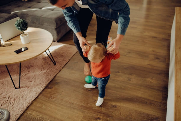 Parent helping a toddler walk on wood-look flooring in living room. -flooring contractors